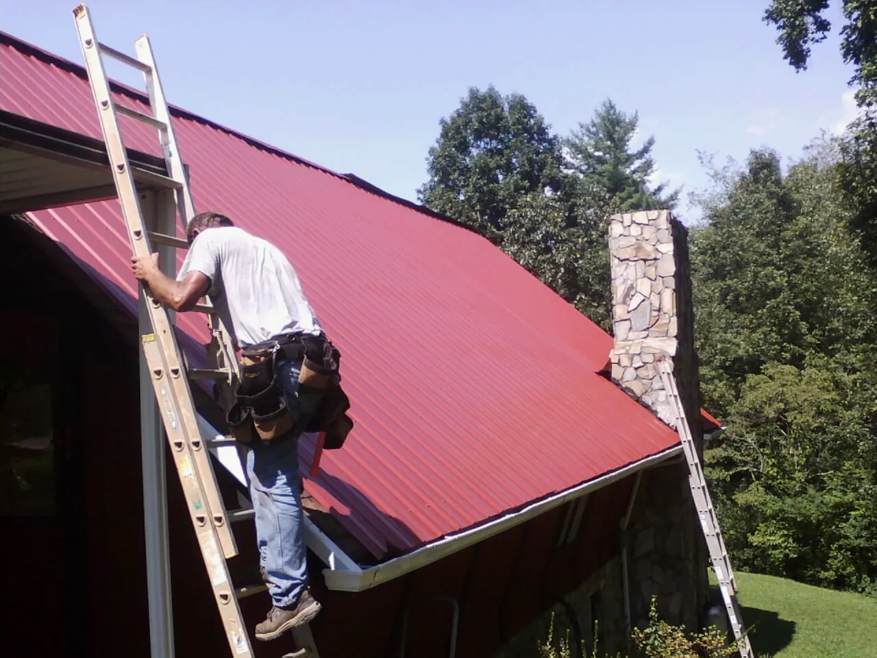 Roofer on a ladder beside a red metal roof and stone chimney — Soffit & Fascia Repair by Roofing Brothers in Newcomb